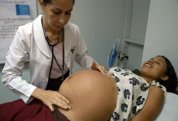 Médica con bata blanca y estetoscopio revisa el vientre de una mujer embarazada recostada en una camilla durante una consulta prenatal.