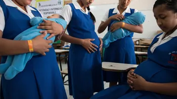 Estudiantes de secundaria con uniforme azul cargan bebés envueltos en cobijas y muestran vientre de embarazo durante una clase de maternidad y paternidad responsable.
